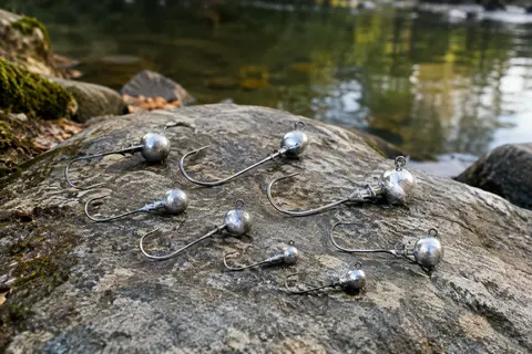 Têtes plombées argentées avec hameçons inox posées sur une pierre au bord d’une rivière calme.