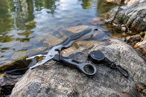 Pince de pêche multifonction grise et noire avec lames dentelées et rétracteur, posée près de l’eau