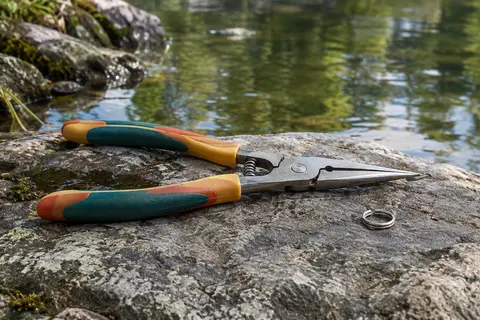 Pince à anneaux brisés à poignées multicolores et bec fin en acier, posée de profil sur une pierre au bord d’une rivière, avec un petit anneau brisé à côté.