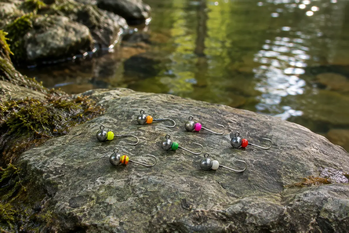 Petites têtes plombées en tungstène aux perles vives et hameçons argentés, posées au bord de l’eau